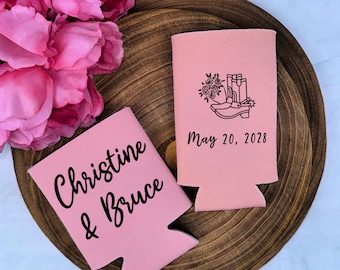 May include: Two light pink foam can coolers. One features the black text "Christine & Bruce" in a script font. The other displays a black line art graphic of cowboy boots with flowers and the black text "May 20, 2028" in a sans-serif font.
