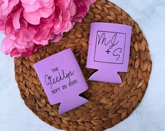 May include: Two lavender can coolers with black text and a woven coaster. One cooler reads "The Stecklys Sept 20 2025". The other cooler has the initials "MJ + S" inside a square. Pink flowers are in the background.