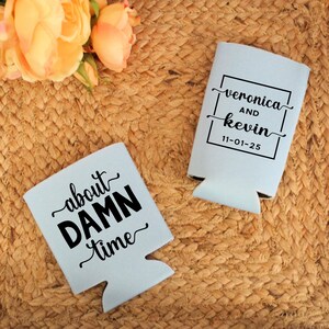 May include: Two light blue can coolers with black text. One says "about DAMN time" and the other says "veronica and kevin" with the date "11-01-25". The coolers are on a woven, tan surface.