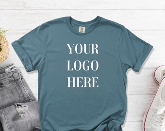 May include: A teal-colored t-shirt with rolled-up sleeves. The text "YOUR LOGO HERE" is printed in white block letters. The shirt is displayed with a pair of gray jeans and white sneakers.