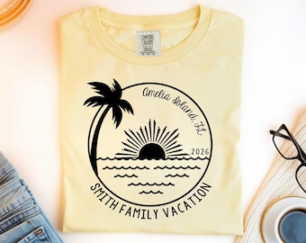 May include: A light yellow t-shirt with a black graphic of a palm tree, sun, and ocean waves. Text reads "Amelia Island, FL 2026" and "Smith Family Vacation" in a circular design. The shirt is folded.