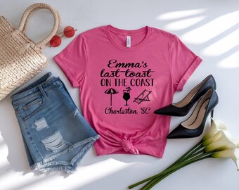 May include: A pink t-shirt with the text "Emma's last toast on the coast" and beach-themed graphics. The shirt is paired with denim shorts, black heels, a straw bag, and sunglasses. The text also includes "Charleston, SC".