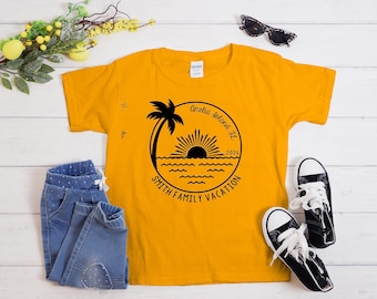 May include: Mustard yellow t-shirt with a black graphic of a palm tree, sun, and waves, with the text "Amelia Island, FL 2026 SMITH FAMILY VACATION". The shirt is paired with blue jeans and black sneakers.
