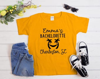 May include: Mustard yellow t-shirt with the text "Emma's Bachelorette Charleston, SC" in black script. The shirt features a smiling palm tree graphic. The image includes blue denim pants, black and white sneakers, and sunglasses.