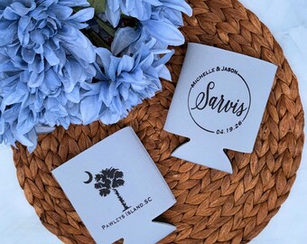 May include: Two gray drink koozies with black text and graphics sit on a woven mat, next to blue flowers. One koozie has a palm tree and the text "PAWLEYS ISLAND. SC". The other has the names "MICHELLE & JASON" and the last name "Sarvis" with the date "04.19.26".