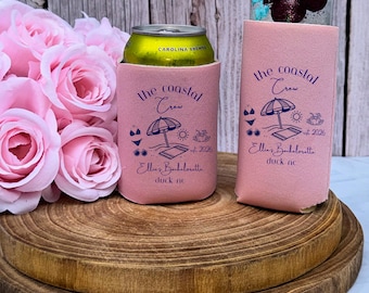 May include: Two light pink foam can coolers featuring dark blue text and graphics. The design includes the words the coastal crew, est 2026, Ellie's Bachelorette, and duck nc, alongside icons of a beach umbrella, sun, bikini, and sunglasses.