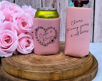 May include: Two pink foam can coolers. One cooler holds a yellow beverage can and features a black floral heart design with the text "Cara & Kevin". The other cooler is tall and features the black text "Cheers to many years & cold beers".