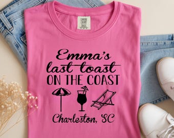 May include: A pink t-shirt with the text "Emma's last toast ON THE COAST" in black script. Below the text are illustrations of a beach umbrella, a cocktail, and a lounge chair. The text "Charleston, SC" is printed below the illustrations.