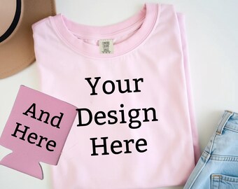 May include: A light pink t-shirt with the text "Your Design Here" in black. A pink can cooler with the text "And Here" is in the foreground. A tan hat and blue jeans are also visible.