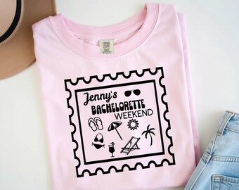 May include: A light pink t-shirt with a black graphic that reads "Jenny's Bachelorette Weekend." The graphic includes sunglasses, sandals, a bikini, an umbrella, a sun, a palm tree, a cocktail, and a beach chair.