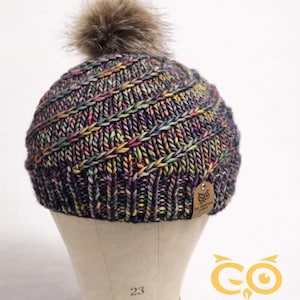 May include: A hand-knitted beanie hat with a multicolored yarn pattern in shades of purple, blue, yellow, and pink. The hat features a ribbed brim and a fluffy, brown faux fur pom-pom on top. A small tag is attached to the hat.