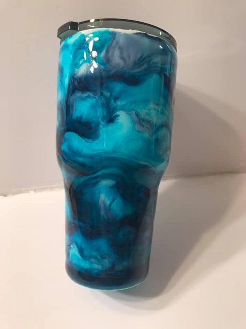 Puede incluir: Un vaso azul y blanco marmoleado con tapa negra. El vaso tiene un patr&oacute;n de remolino de azul, blanco y negro.