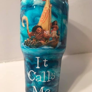 Puede incluir: Un vaso azul y blanco con un patr&oacute;n de remolino. Presenta una imagen de Maui y Moana de la pel&iacute;cula de Disney Moana, navegando en un barco. El texto "It Calls Me" est&aacute; impreso en el vaso.