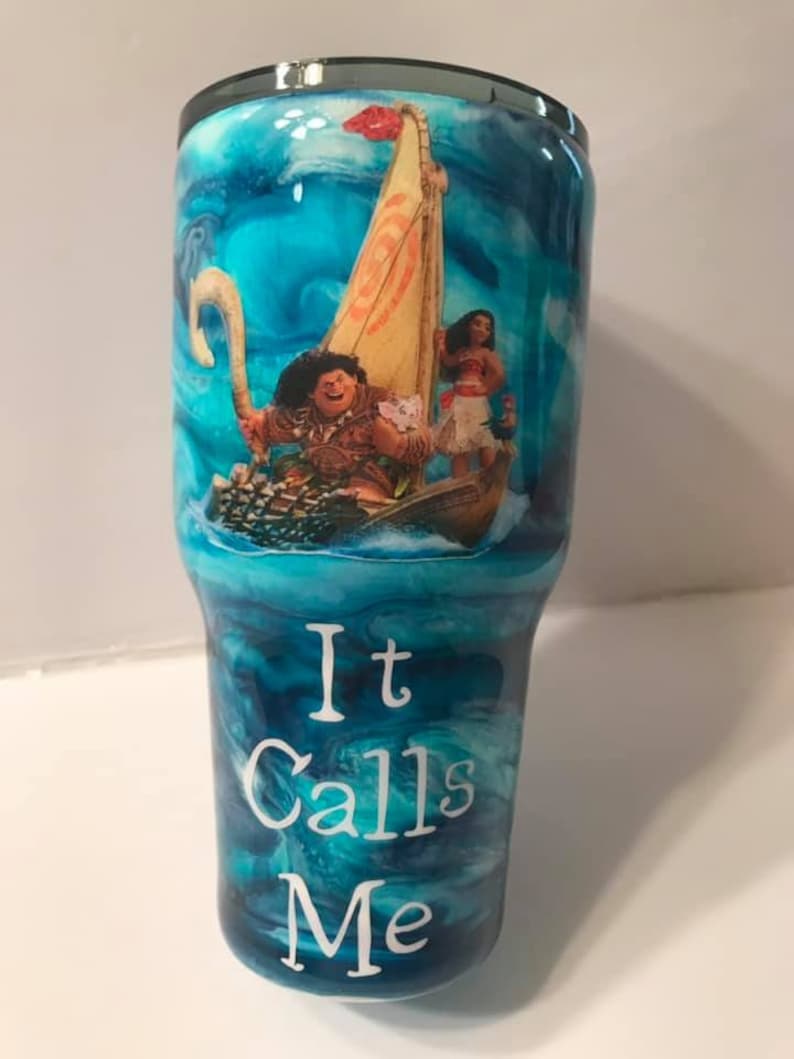 Puede incluir: Un vaso azul y blanco con un patr&oacute;n de remolino. Presenta una imagen de Maui y Moana de la pel&iacute;cula de Disney Moana, navegando en un barco. El texto "It Calls Me" est&aacute; impreso en el vaso.
