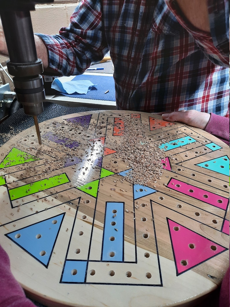 May include: A person using a drill press to create holes in a wooden board. The board is round and features a colorful geometric design with blue, green, and pink shapes. Sawdust is visible around the drill.