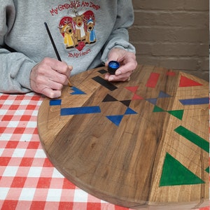 May include: A person painting a wooden, heart-shaped cutting board with geometric designs. The board features blue, red, black, and green shapes. The person is wearing a gray sweatshirt with the text "My Grandkids Are Dear To My Heart".