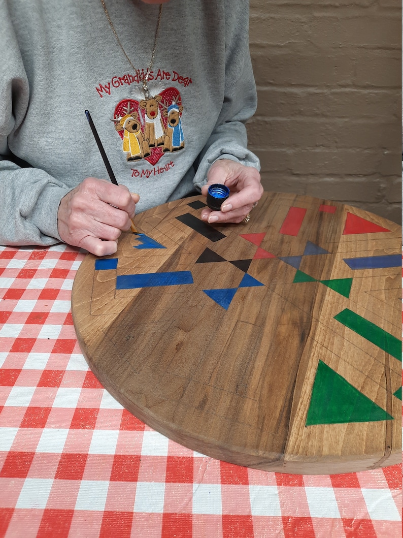 May include: A person painting a wooden, heart-shaped cutting board with geometric designs. The board features blue, red, black, and green shapes. The person is wearing a gray sweatshirt with the text "My Grandkids Are Dear To My Heart".