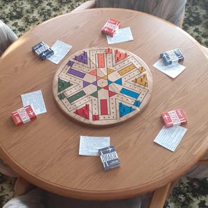 May include: A wooden cribbage board with colorful pegs and a starburst design sits on a round wooden table. Small boxes of matches with the word "AVIATOR" are scattered around the board, along with small pieces of paper.