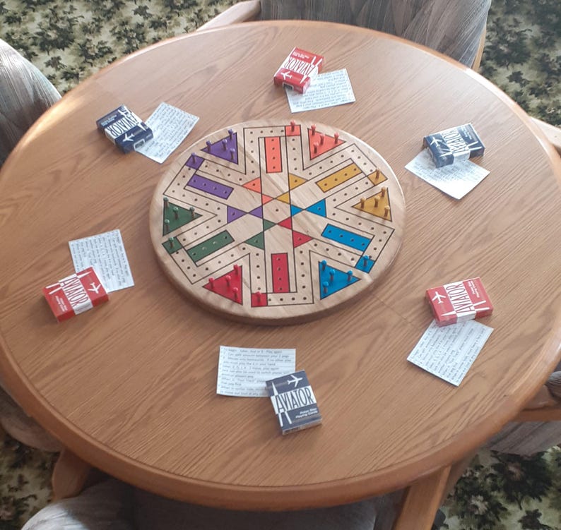 May include: A wooden cribbage board with colorful pegs and a starburst design sits on a round wooden table. Small boxes of matches with the word "AVIATOR" are scattered around the board, along with small pieces of paper.