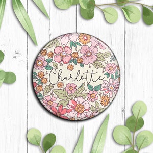 May include: A round button featuring a floral design in shades of pink, yellow, and green. The name "Charlotte" is written in black cursive script in the centre of the button.