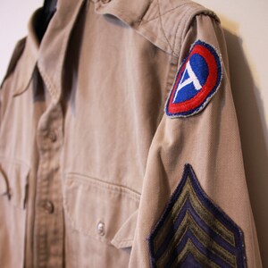 Puede incluir: Una camisa militar de color beige con un parche azul y rojo en la manga derecha y un parche morado y verde en la manga izquierda. Los parches son insignias militares.