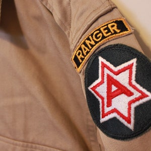 Puede incluir: Una chaqueta militar de color beige con un parche negro y rojo que dice "Ranger" y un parche verde con una estrella roja y la letra "A" en su interior.