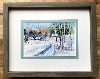May include: A watercolor painting of a snowy winter scene with two small houses and a winding road leading into a forest. The trees are bare and the snow is white and fluffy. The painting is framed in a gray frame with a light blue mat.