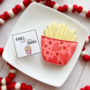May include: A red and white cookie decorated with a heart pattern and a drawing of french fries. The cookie is on a white plate with a white card that says "Fries before guys".