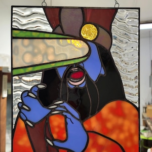 May include: Stained glass panel featuring a stylized figure with blue skin, black hair, and an open mouth. The figure has blue hands with long white nails. The panel incorporates red, orange, yellow, and green glass, with a black metal frame and hanging chain.