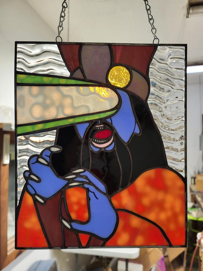 May include: Stained glass panel featuring a stylized figure with blue skin, black hair, and an open mouth. The figure has blue hands with long white nails. The panel incorporates red, orange, yellow, and green glass, with a black metal frame and hanging chain.