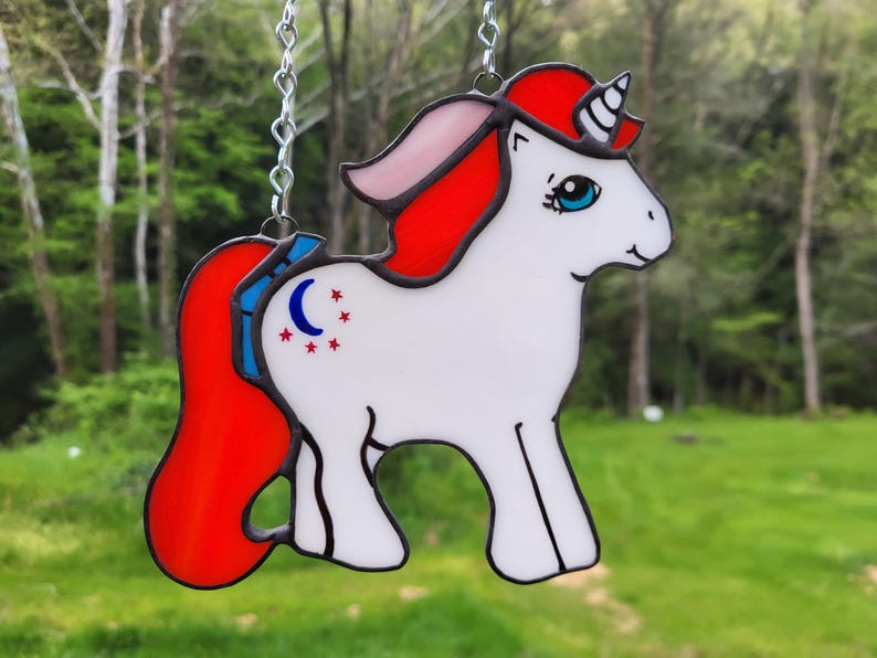Pu&ograve; includere: Un unicorno in vetro colorato con criniera e coda rosse, corpo bianco e una mezzaluna blu e argento e stelle sul fianco.