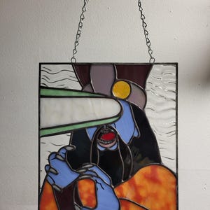 May include: Stained glass panel featuring an abstract design with a figure in blue, black, and orange. The figure has a red mouth and is holding a white object. The panel is framed in black and suspended by a silver chain.