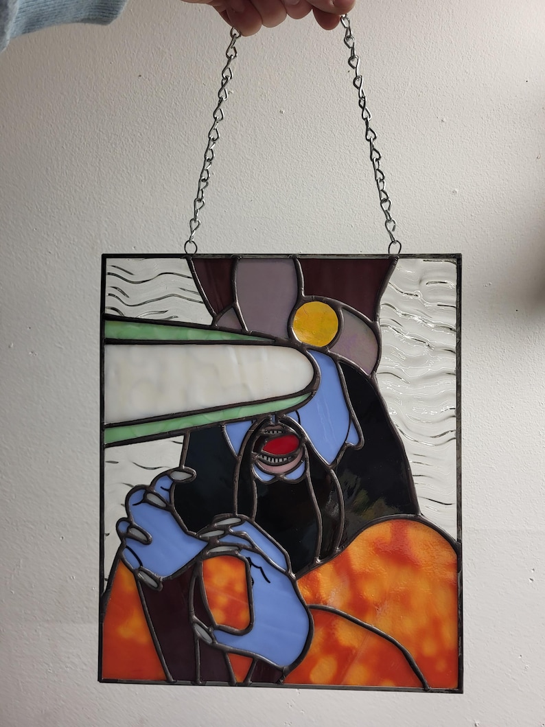 May include: Stained glass panel featuring an abstract design with a figure in blue, black, and orange. The figure has a red mouth and is holding a white object. The panel is framed in black and suspended by a silver chain.