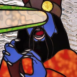 May include: Stained glass artwork featuring a stylized figure with blue skin, black hair, and an open mouth. The figure has blue hands and is wearing an orange and red garment. A green and white shape is above the figure's head. The artwork is signed "sunspotsbyadria.com".