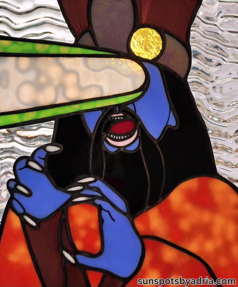 May include: Stained glass artwork featuring a stylized figure with blue skin, black hair, and an open mouth. The figure has blue hands and is wearing an orange and red garment. A green and white shape is above the figure's head. The artwork is signed "sunspotsbyadria.com".