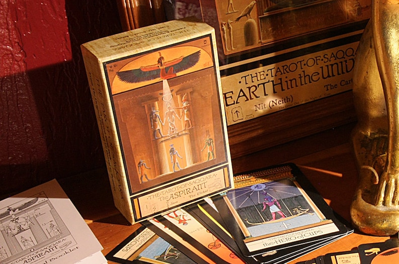 May include: A box of tarot cards with an Egyptian theme. The box features a golden background with a winged figure and hieroglyphics. The cards are spread out on a wooden surface. The text on the box reads "The Tarot of Saqqara, The Aspirant".
