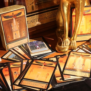 May include: A deck of tarot cards with Egyptian-themed imagery, including a card with a winged sun god and the text "THE TAROT OF SAQQ/ EARTH Hintheuniv". The cards are spread out on a dark surface with a gold statue in the background.