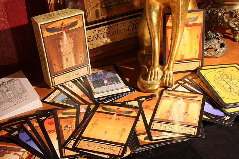 May include: A deck of tarot cards with Egyptian-themed imagery, including a card with a winged sun god and the text "THE TAROT OF SAQQ/ EARTH Hintheuniv". The cards are spread out on a dark surface with a gold statue in the background.