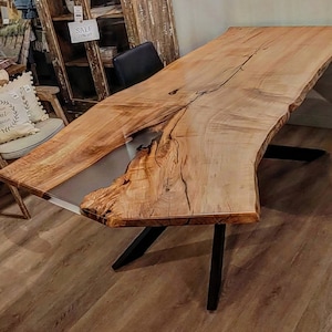 Peut inclure: Une grande table en bois à bord vif avec une finition en bois naturel et une rivière d'époxy transparente traversant le centre. La table a un piètement en X en métal noir.