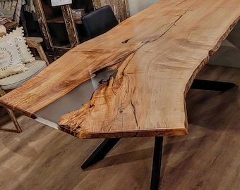 Może przedstawiać: Duży, żywy stół drewniany z naturalnym wykończeniem drewna i przezroczystą rzeką epoksydową biegnącą przez środek. Stół ma czarną metalową podstawę w kształcie litery X.