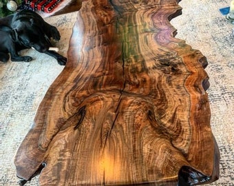 May include: A large, live edge coffee table with a dark wood grain and a natural, irregular shape. The table has a black base and is sitting on a white rug.