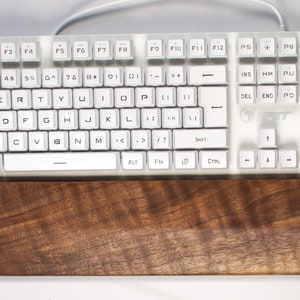 Op de afbeelding: Een wit toetsenbord met een polsrust van walnoot hout. Het toetsenbord heeft een standaard lay-out met functietoetsen, cijfertoetsen en een numeriek toetsenblok. De polsrust is van natuurlijke houtkleur met een nerfpatroon.