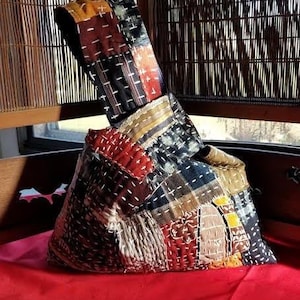 May include: A patchwork bag with a unique design. The bag features a variety of fabric patches in shades of red, brown, black, and cream, stitched together with white thread. The bag is set on a red surface.