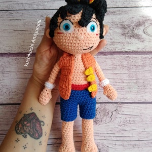 May include: A handmade crochet doll featuring a character with black hair, blue eyes, and a peach skin tone. The doll wears an orange vest, blue shorts, and white wristbands. The doll is approximately 10 inches tall.