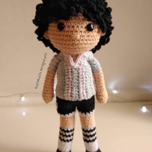 Puede incluir: Muñeca de ganchillo hecha a mano con cabello rizado negro, que lleva una camisa azul claro y gris, pantalones cortos negros y calcetines a rayas. La muñeca tiene un tono de piel bronceado y ojos negros. Creación de estilo amigurumi.