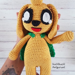 May include: Crocheted yellow amigurumi doll with large white eyes, black accents, and a red mouth. The doll has a green cape and a tall, pointed head. The doll is handmade and has the text "Kuchikuchi Amigurumi" on the side.