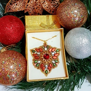 Può includere: Una collana con un ciondolo a forma di fiocco di neve in perle dorate, rosse, verdi e bianche in una scatola regalo. Il fiocco di neve è circondato da una catena dorata.