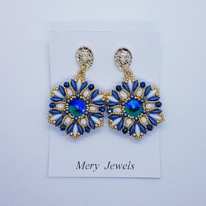 Può includere: Un paio di orecchini con perline blu e bianche con dettagli dorati e un cristallo blu al centro. Gli orecchini sono appesi a una carta bianca con il testo "Mery Jewels".