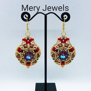 May include: A pair of gold, red, and brown beaded earrings with a blue crystal in the center. The earrings are hanging from a black stand. The text "Mery Jewels" is visible at the top of the image.