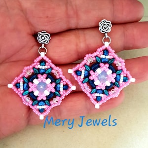 May include: Pink and blue beaded earrings with a white center and silver rose-shaped earring hooks. The earrings are square shaped and have a geometric design. "Mery Jewels" is written in blue text below the earrings.